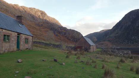 A-slow-right-pan-reveals-multiple-abandoned-old-stone-buildings-and-a-Scottish-Bothy-called-Glendhu-set-amongst-steep-cliff,-mountains-and-a-sea-loch-in-a-remote-glen-in-rural-Scotland