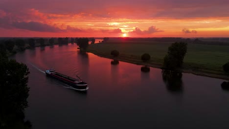 Eine-Luftaufnahme-Eines-Wunderschönen-Sonnenuntergangs-über-Dem-Holländischen-Fluss-Mit-Einem-Vorbeifahrenden-Schiff-In-Einer-Orangefarbenen-Und-Roten-Landschaft