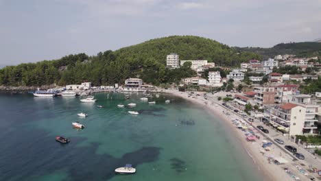 Pullback-from-Himare-turquoise-Water-bay-with-small-boat-pier,-the-sand-beach-coastline,-Albania