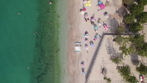 Topdown-ascending-view-of-Beach-shoreline-with-emerald-colored-water,-Colorful-sun-hats-in-white-sand-beach,-Himare