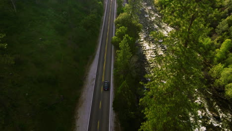 Drone-Aerial-Following-A-Blue-Tesla-Car-Driving-On-The-Road-Next-To-The-River-With-Lush-Vegetation-Near-Yosemite-National-Park-In-California,-USA