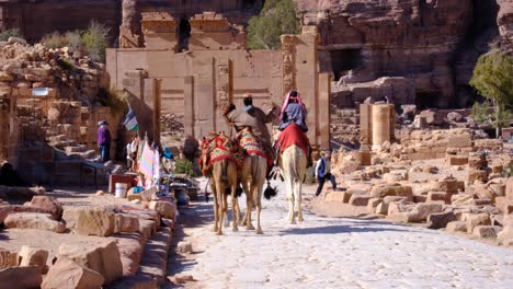 Arabian-people-riding-camels-on-rocky-pathway-lined-with-market-stalls-and-ancient-tombs-in-the-distance-in-the-city-of-Petra,-Jordan,-Middle-East