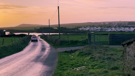 Am-Frühen-Morgen-Im-Winter-Mit-Aufgehender-Sonne-über-Dem-Ozean-Und-Dem-Auto,-Das-Auf-Der-Landstraße-In-Irland-Vorbeifährt