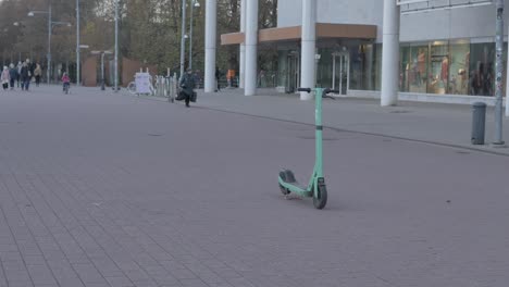 La-Gente-Está-Caminando-En-El-Fondo-En-Cámara-Lenta,-Detrás-De-Un-Scooter-Eléctrico-De-Perno-Verde