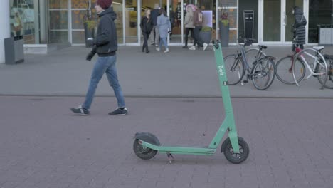 Die-Leute-Gehen-In-Zeitlupe-Des-Grünen-Bolzen-elektrorollers-Vorbei,-Der-Bolzen-Wurde-Mitten-In-Der-Fußgängerzone-Geparkt