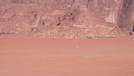 White-4WD-truck-driving-across-vast,-remote-Wadi-Rum-desert-in-Jordan-with-Bedouin-tents-nestled-against-mountains-in-the-distance