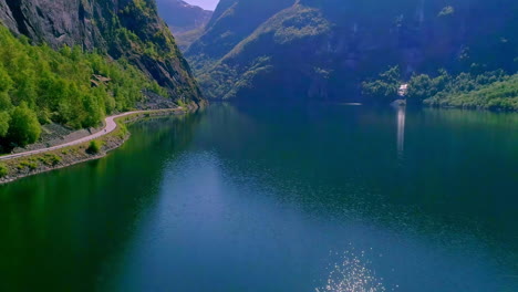 Atemberaubende-Luftaufnahme-Eines-Sees,-Der-Im-Sommer-Von-Riesigen-Bergen-Mit-Einer-Kleinen-Straße-Am-Wasser-Beschallt-Wird