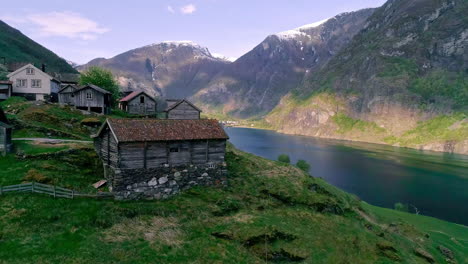 Aerial-view-of-old-wooden-houses-at-Fjord-of-Norway-with-snowy-mountaintops-in-background-in-summer