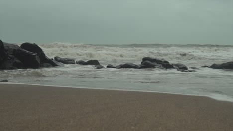 Wellen,-Die-Auf-Eine-Felsige-Küste-Krachen-Und-Sand-Zurückziehen,-Während-Das-Wasser-Zurück-Ins-Meer-Fließt