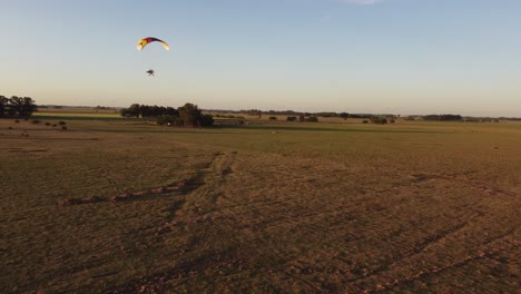 Aerial-tracking-shot-of-Motorized-paratrike-paraglider-flying-over-rural-field-at-golden-sunset