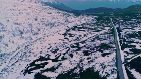 Aerial-flyover-snowy-winter-landscape-in-Norway-with-road-and-iced-mountains-range-in-background-during-sunset