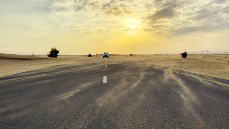 Ein-Weißer-Geländewagen-Fährt-Auf-Einer-Straße-Mitten-In-Der-Wüste,-Während-Der-Wind-Bei-Sonnenuntergang-Die-Feinen-Sandpartikel-Verweht