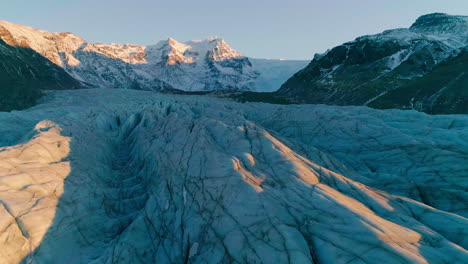 Skaftafell-Glacier-sunlit-curving-Vatnajökull-national-park-glacier-tilt-up-to-mountain-range,-Aerial-view