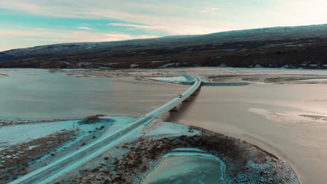 Aerial-view-orbiting-Múlaþing-Egilsstaðir-West-Fjords-bridge-crossing-estuary-to-colourful-sunset-mountain,-Iceland