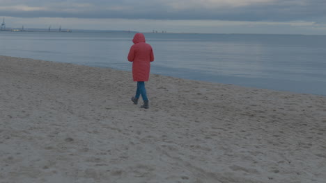 Person-with-orange-raincoat-walking-on-sandy-beach-beside-ocean-during-cloudy-day
