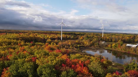 Wind-turbines-surrounded-by-colorful-autumn-forest-and-lakes,-aerial-fly-forward-view