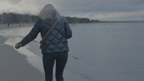 View-From-Behind-Women-With-Blond-Hair-Holding-Dog-Lease-Walking-Along-Beach-In-Gdansk,-Poland