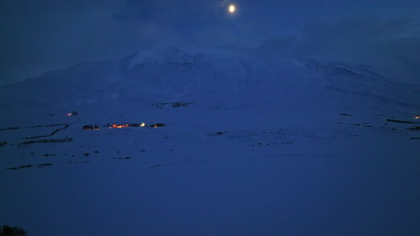 Luz-Brillante-Sobre-Una-Montaña-Nevada-En-El-Paisaje-Invernal-Con-Iluminación-Esporádica-De-Casas-De-Islandia-Durante-La-Noche