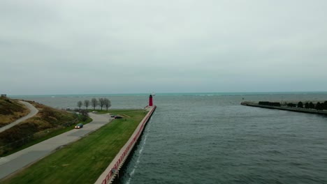 Aerial-shot-over-a-river-to-a-red-lighthouse-on-a-cloudy-day,-sea-in-the-background