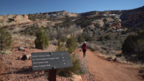 Colorado-National-Monument-Schild-Mit-Wegbeschreibungen-Zum-Wanderweg-Und-Rücken-Einer-Frau,-Die-Auf-Dem-Weg-Geht