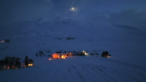 Luz-Brillante-Sobre-Una-Montaña-Nevada-En-El-Paisaje-Invernal-Con-Iluminación-Esporádica-De-Casas-De-Islandia-Durante-La-Noche