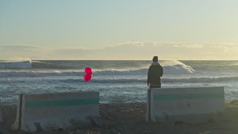A-man-holds-three-red-balloons-as-he-watches-the-ocean-waves-break-before-him