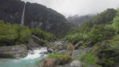 Mujer-Aventurera-Caminando-En-La-Orilla-Del-Río-Glacial-Deteniéndose-Para-Tomar-Una-Foto,-Glaciar-Rob-Roy