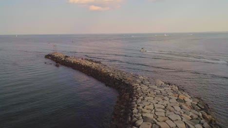 Rocky-path-into-the-ocean,-with-boats-going-by--drone-video