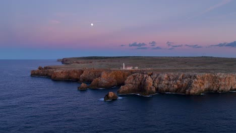 Distant-aerial-orbit-around-Punta-Nati-Lighthouse-on-rocky-cliffs-of-Menorca,-Spain