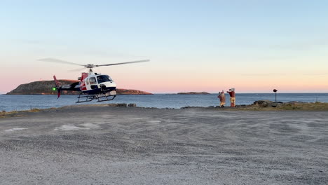 Plano-Medio-De-Un-Helicóptero-Despegando-Cerca-De-Un-Cuerpo-De-Agua-Durante-La-Puesta-De-Sol-Con-Una-Isla-Rocosa-En-El-Fondo