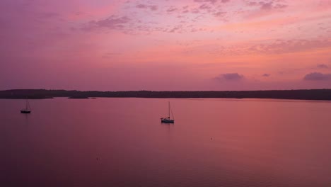 Aerial-view-of-two-boats-in-the-ocean-in-Menorca-Spain-during-a-pink,-purple,-orange-sunrise,-tracking-wide-shot