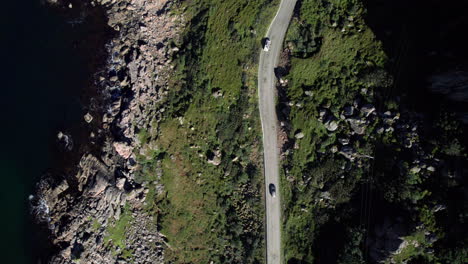 Aerial-birdseye-view-of-a-car-driving-along-a-curvy-highway-on-the-hilly-and-rocky-coast-on-a-bright-summer-day