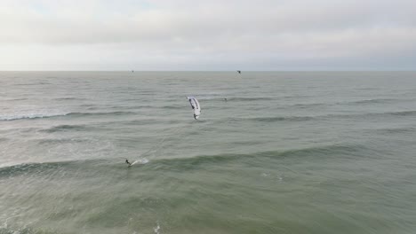 Establecimiento-De-Una-Vista-Aérea-De-Un-Grupo-De-Personas-Dedicadas-Al-Kitesurf,-Día-De-Invierno-Nublado,-Olas-Altas,-Deporte-Extremo,-Playa-De-Karosta-Del-Mar-Báltico,-Amplio-Tiro-De-Drones-Moviéndose-Hacia-Atrás