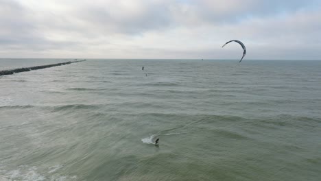 Establecimiento-De-Una-Vista-Aérea-De-Un-Grupo-De-Personas-Dedicadas-Al-Kitesurf,-Día-De-Invierno-Nublado,-Olas-Altas,-Deporte-Extremo,-Playa-De-Karosta-Del-Mar-Báltico,-Tiro-Panorámico-De-Drones