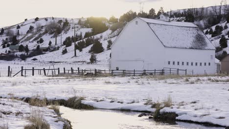 Weiße-Scheune-Auf-Schneebedeckter-Weide-Mit-Schönem-Fluss,-Der-Durchfließt,-Winterlandschaft,-Ranching-Lebensstil,-4k