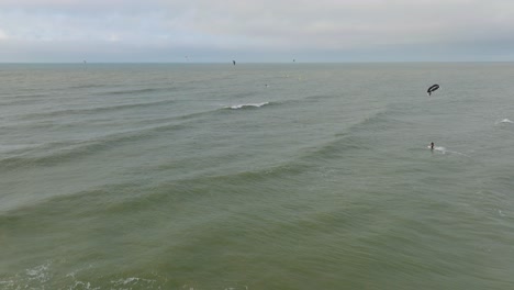 Establishing-aerial-view-of-a-group-of-people-engaged-in-kitesurfing,-overcast-winter-day,-high-waves,-extreme-sport,-Baltic-Sea-Karosta-beach-,-wide-drone-shot-moving-forward