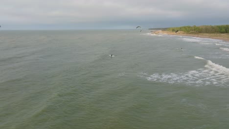 Erstellung-Einer-Luftaufnahme-Einer-Gruppe-Von-Menschen,-Die-Kitesurfen-Betreiben,-Bewölkter-Wintertag,-Hohe-Wellen,-Extremsport,-Ostsee-Karosta-Strand,-Entfernter-Drohnenschuss,-Der-Sich-Rückwärts-Bewegt