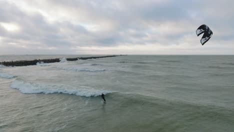 Establishing-aerial-view-of-a-man-engaged-in-kitesurfing,-overcast-winter-day,-high-waves,-extreme-sport,-Baltic-Sea-Karosta-beach-,-drone-shot-moving-backward