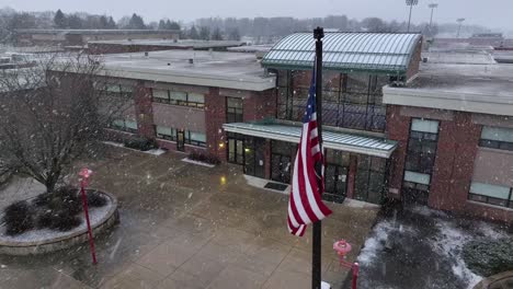 Aerial-orbit-around-American-flag-in-front-of-high-school-building-in-America-on-snow-day