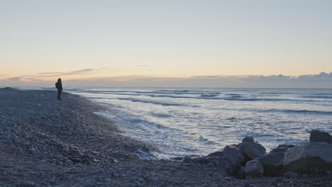 Man-walks-along-stoney-shore-in-early-morning-as-waves-wash-up-onshore