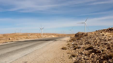 Conducción-De-Automóviles-En-Una-Carretera-Larga-Y-Recta-A-Través-De-Un-Parque-De-Turbinas-Eólicas-En-El-Remoto-Paisaje-Desértico-De-Jordania,-Oriente-Medio