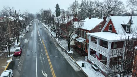 Steigende-Luftaufnahme-Der-Amerikanischen-Flagge,-Die-Von-Der-Veranda-Des-Hauses-Auf-Der-Hauptstraße-Der-Verschneiten-Kleinstadt-In-Amerika-Weht