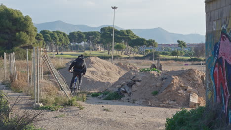 Cyclist-cicles-trough-buildingsite-around-sand-heaps-,-wire-fence-on-one-side-and-wall-with-graffitti-on-other-side,-nice-landscape-with-trees-and-mountains-in-distance