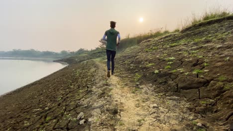 Rear-view-of-man-jogging-by-river,-heavy-drought-in-Bangladesh,-slowmo,-day