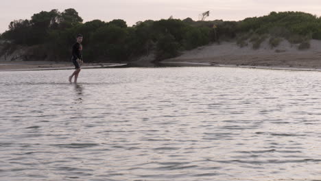 Young-Boy-Walks-Across-Calm-Shallow-River-Water-At-Dusk,-SLOW-MOTION
