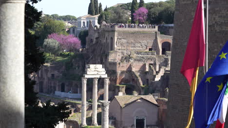 Long-shot-of-the-Roman-Forum-with-tourists-up-on-an-observation-deck-and-the-Roman,-Italian,-and-European-flags-waving-in-the-foreground