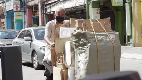 Un-Anciano-Con-Máscara-Empujando-Un-Carro-Lleno-De-Cartón-De-La-Basura-A-Lo-Largo-De-La-Concurrida-Calle-Kuala-Lumpur