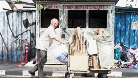 Old-asian-man-pushes-cart-with-collected-cardboard,-garbage-container-in-background