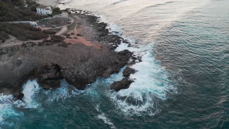 Drone-Top-Down-Shot-Crashing-Waves-at-Shore-Coastline-in-Mallorca-with-blue-water-revealing-the-rest-of-the-island-and-nearby-beach-during-golden-hour