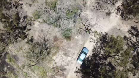 Drone-shot-looking-down-on-blue-car-driving-on-dirt-road-through-trees